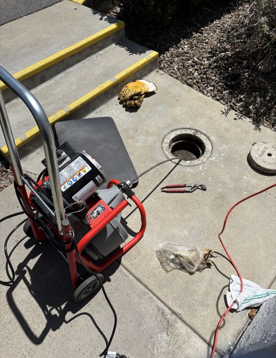 A drain cable feeding into an open sewer cleanout in a concrete walkway, with the concrete cleanout lid set aside.